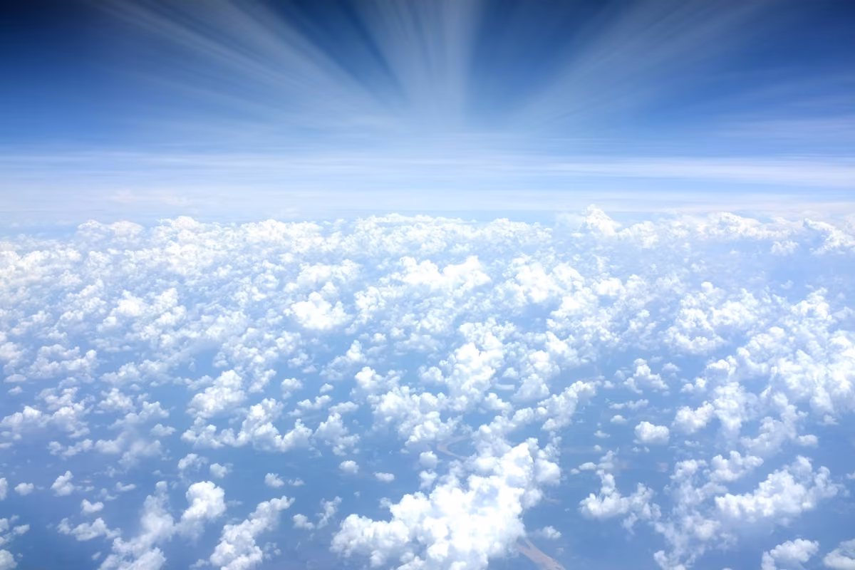 Aerial view of a blanket of white clouds above the horizon with sunbeams piercing through a clear blue sky.