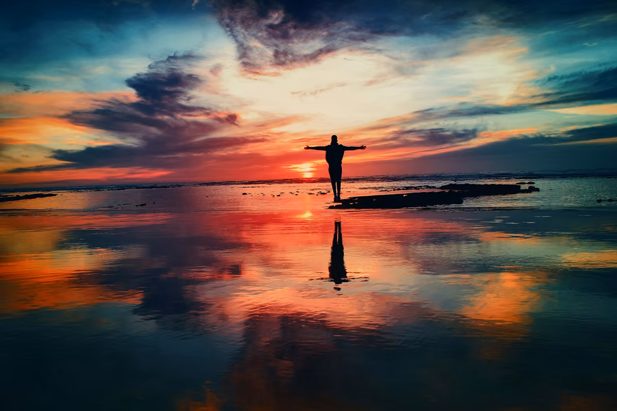 Silhouette of a person standing with open arms facing the sea during a dramatic sunset, reflected on the water.