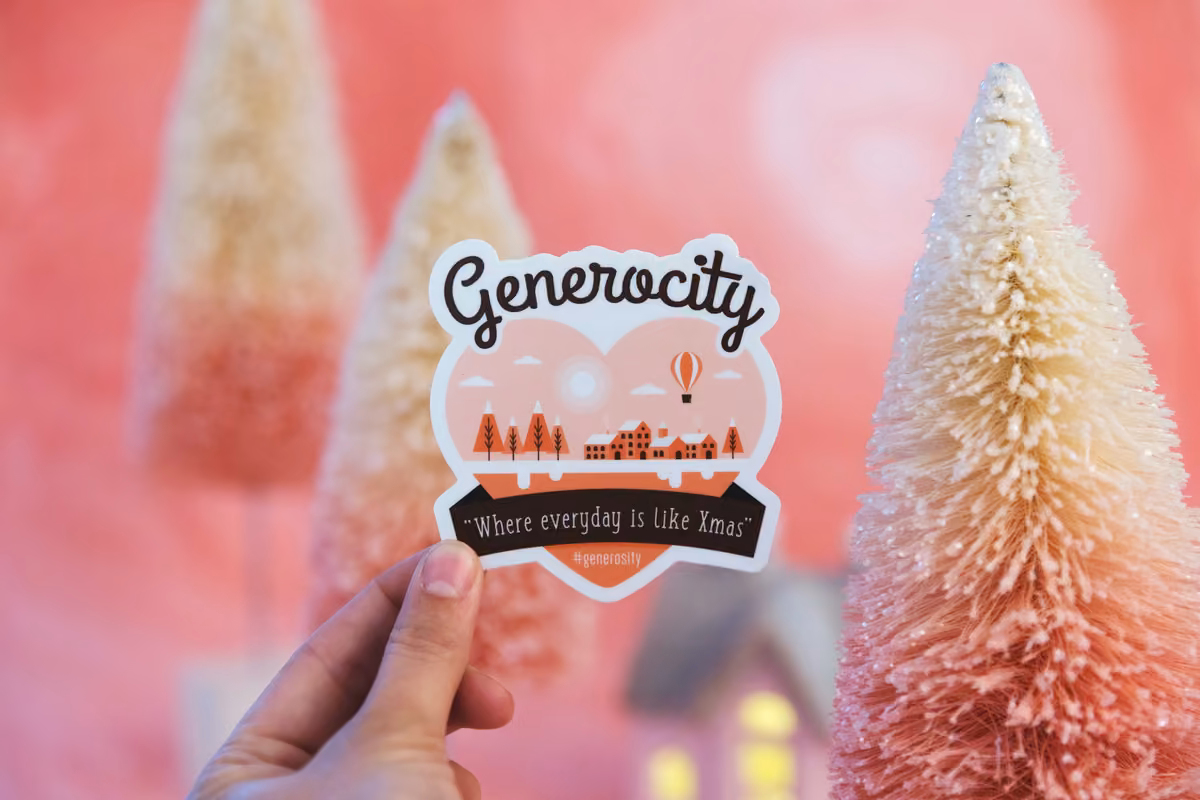 A hand holding an illustrated sticker that reads 'Generocity' in front of a background of pink and white decorative holiday pine trees.