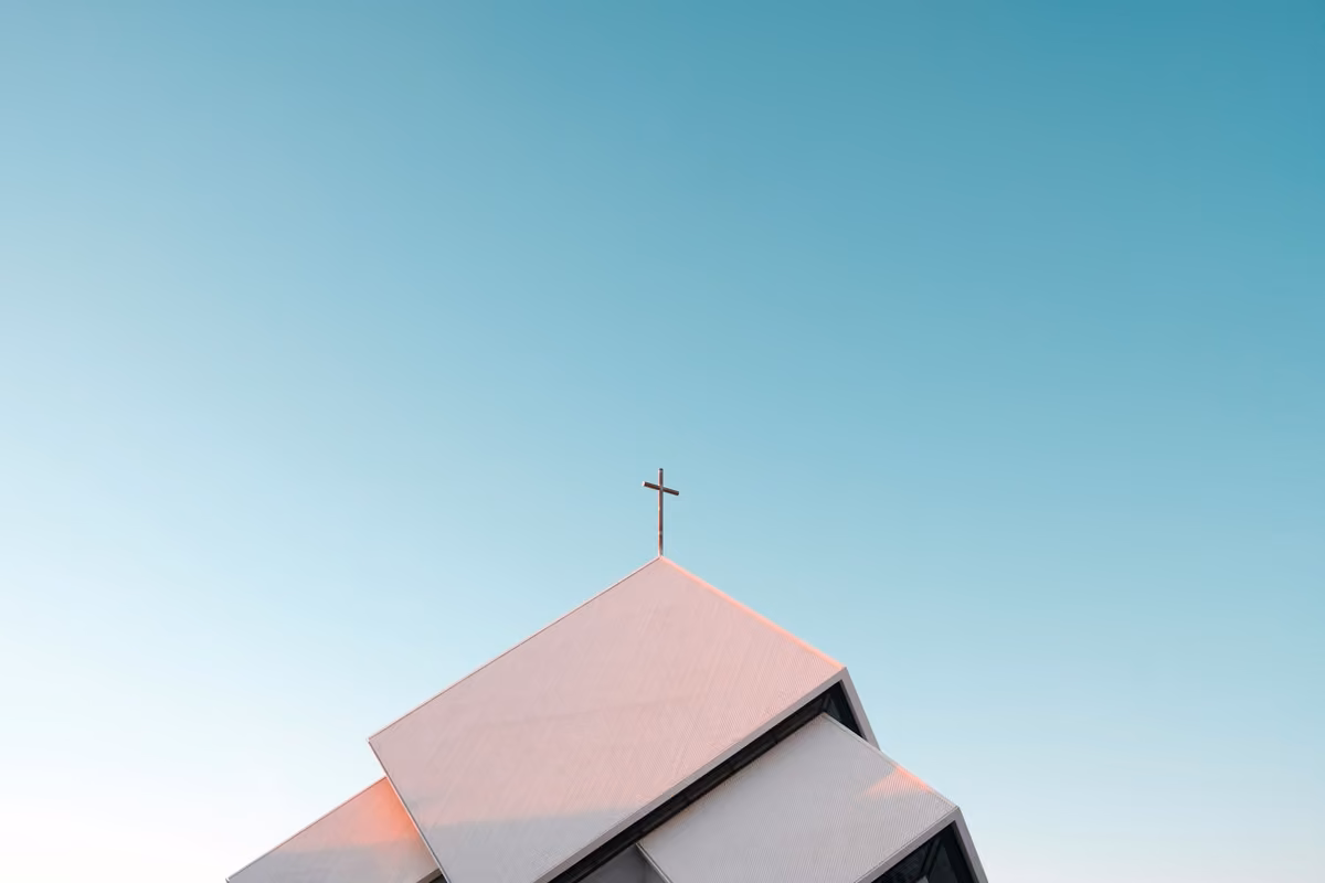 Modern architectural roof of a white church with a cross at the top against a clear blue sky.