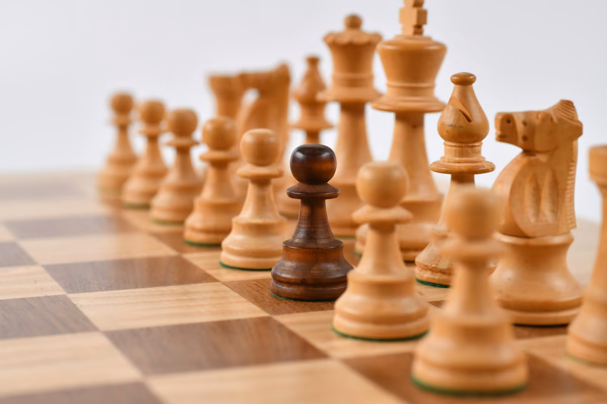 A black chess pawn standing out amidst a row of light-colored wooden chess pieces on a board.