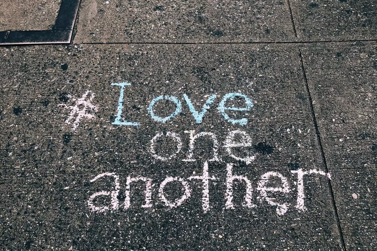 The hashtag '#LoveOneAnother' written in white and blue chalk on a concrete sidewalk.
