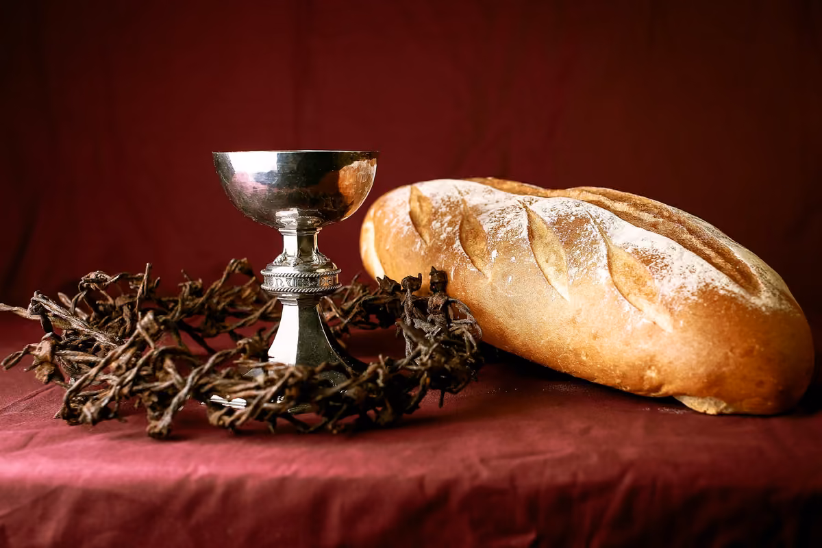 A silver chalice, an artisan loaf of bread, and a crown of thorns on a red tablecloth, representing the symbols of the Lord's Supper.
