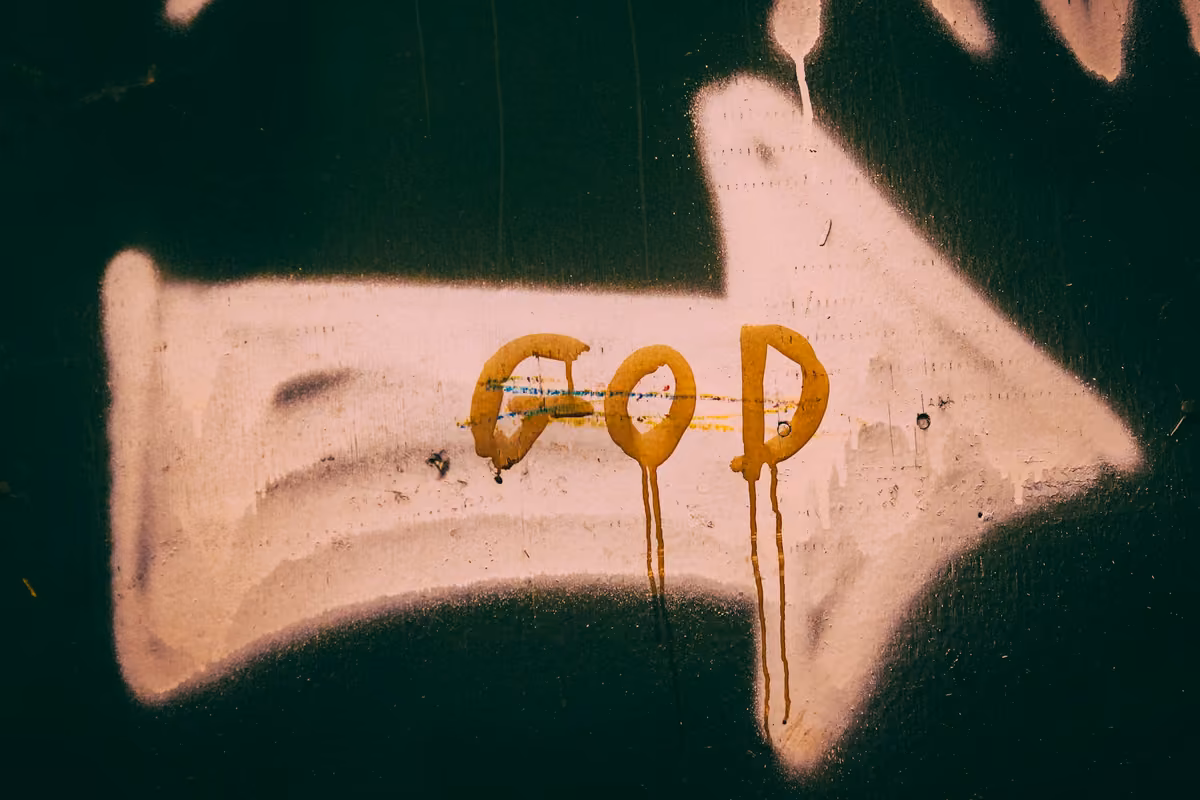 Graffiti of a white arrow on a dark wall with the word «GOD» written in dripping yellow paint.