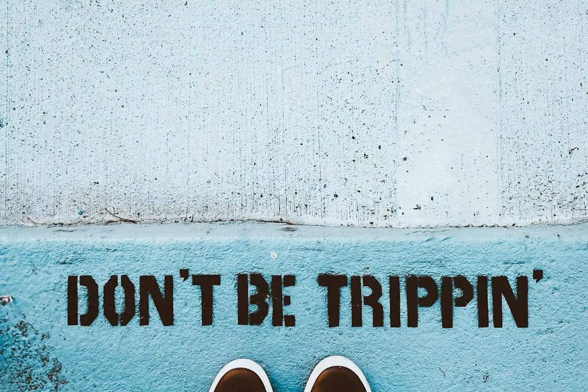 Top-down view of feet in dark sneakers standing in front of the phrase 'DON'T BE TRIPPINT' painted in black on a blue concrete curb.