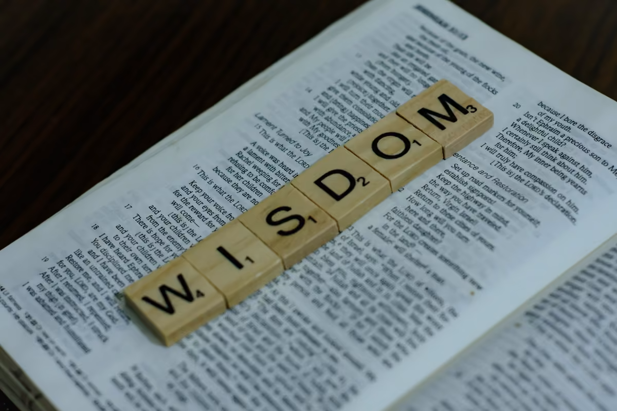 Scrabble-style wooden tiles forming the word 'WISDOM' placed on top of the pages of an open Bible.