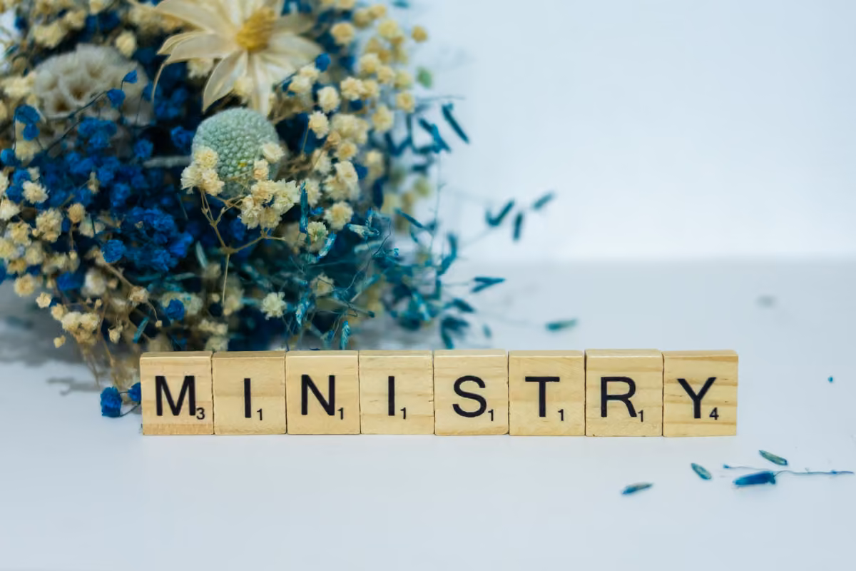 Wooden tiles spelling the word 'MINISTRY' beside an arrangement of dried blue, yellow, and cream flowers on a white surface.