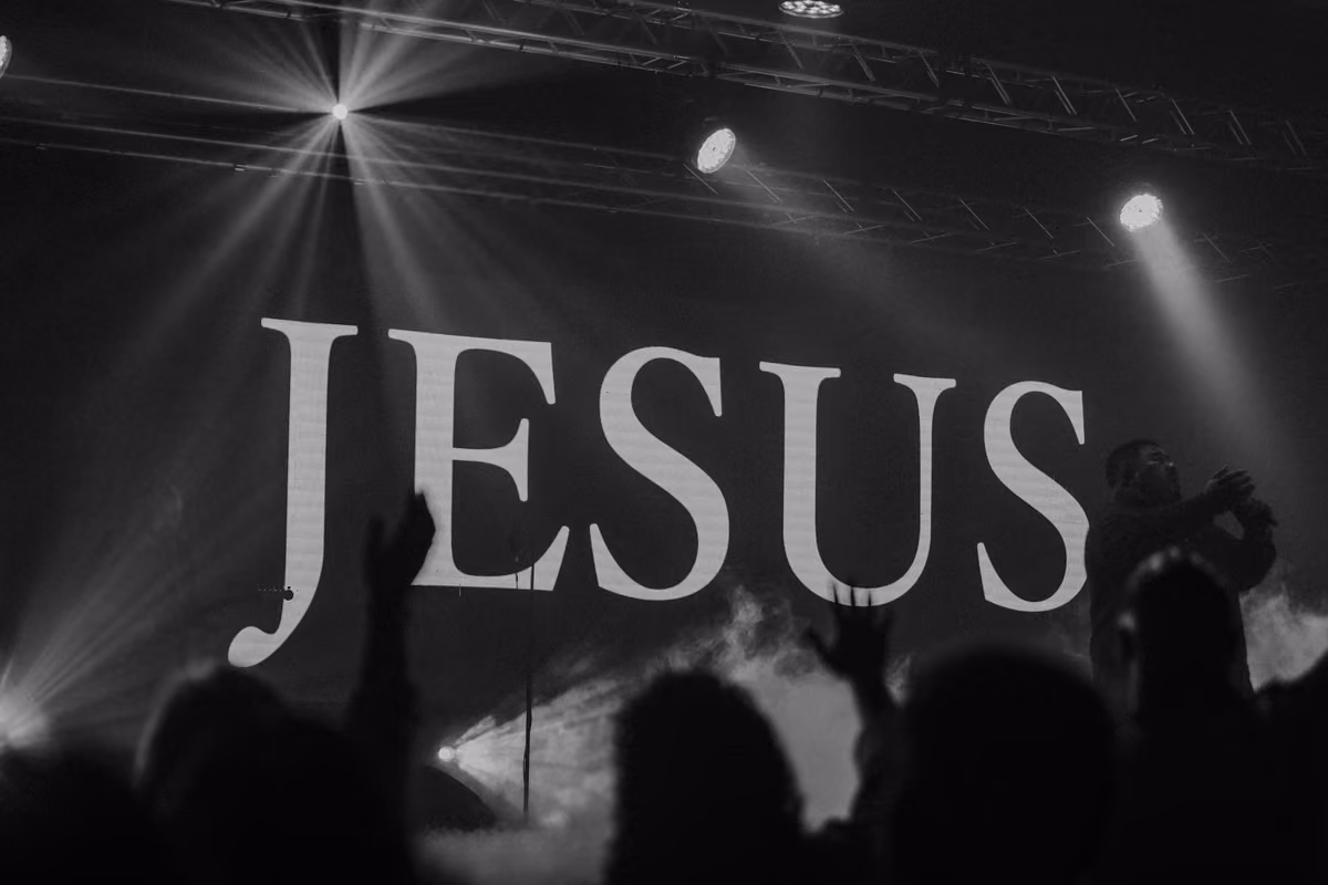 Black and white photograph of a concert or religious service with the word «JESUS» projected on a large screen in the background while the audience raises their hands.