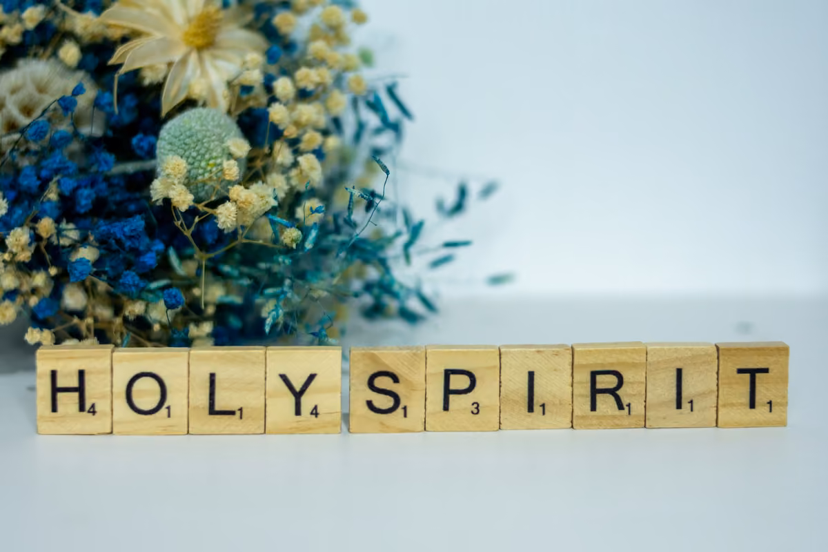 Wooden Scrabble tiles forming the words 'HOLY SPIRIT' next to an arrangement of dried flowers in blue, white, and green tones.