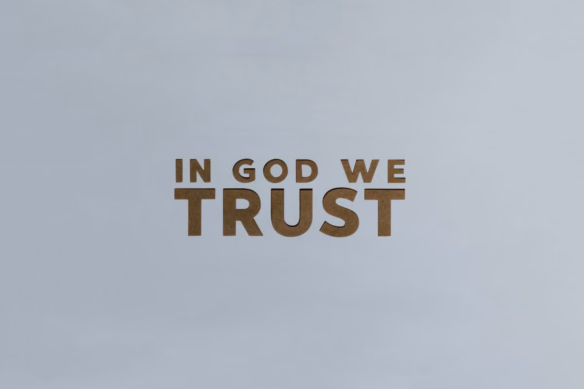 Wooden letters cut out on a light gray background forming the motto 'IN GOD WE TRUST'.