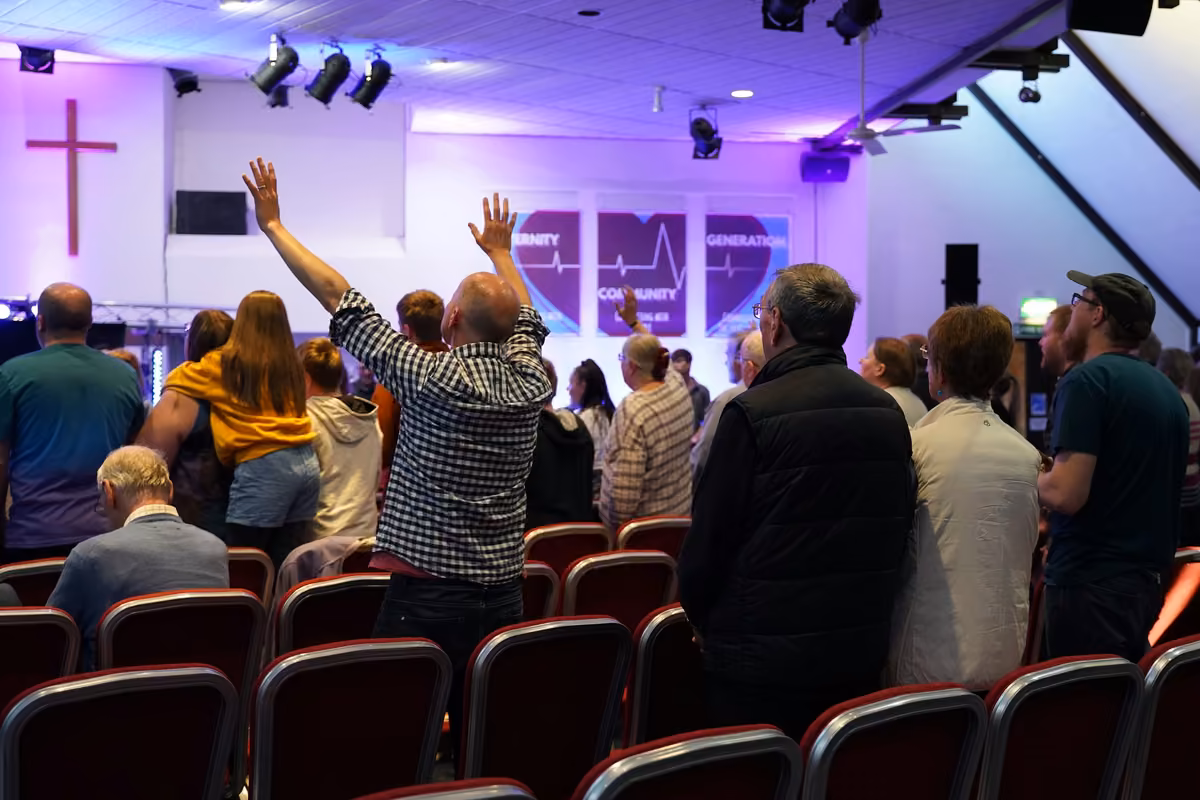 Worshipers standing inside a church illuminated with purple lights, some raising their arms toward a wooden cross on the stage.