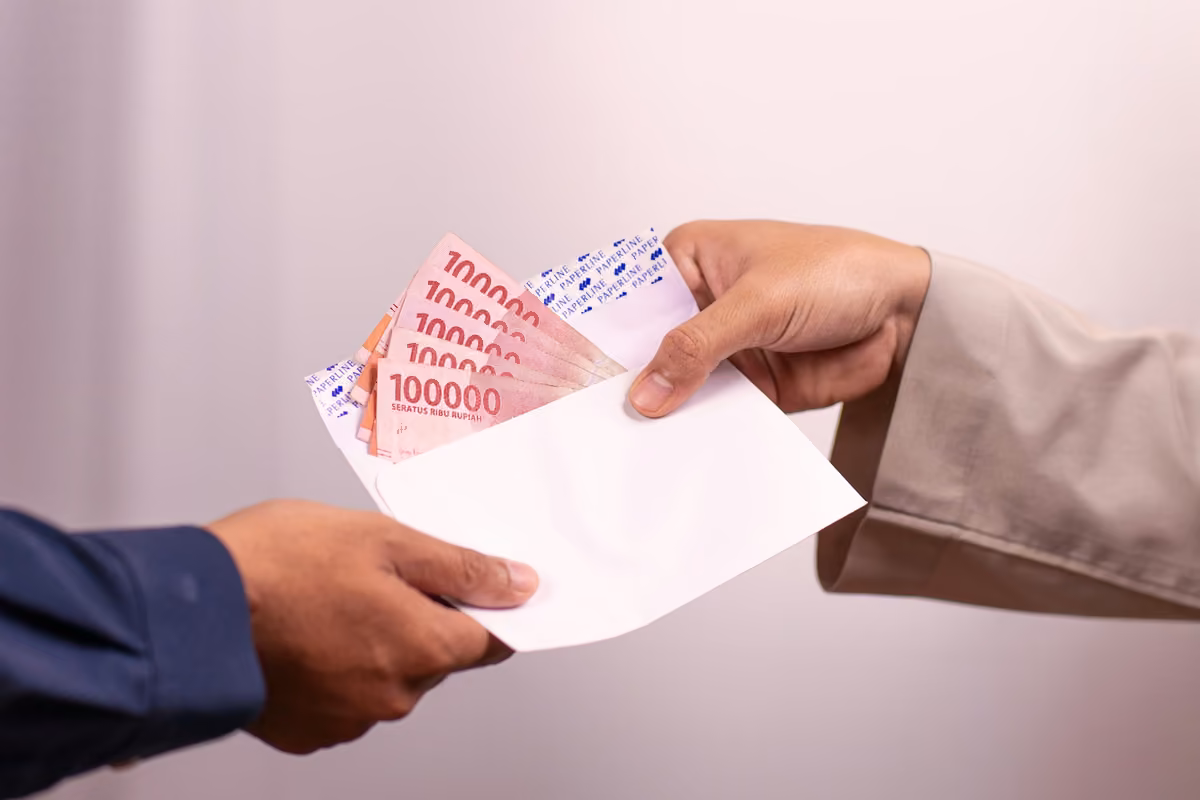 A person handing another an open white envelope filled with 100,000 Indonesian rupiah banknotes.
