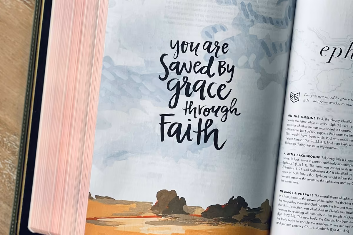 An illustrated page of an open Bible with the calligraphed quote 'You are saved by grace through faith' on a background painted with soft tones.