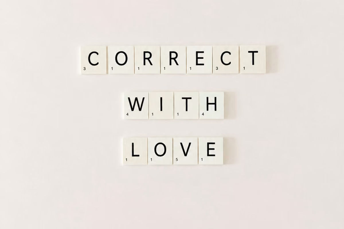 Board game tiles with black letters spelling out the phrase 'CORRECT WITH LOVE' on a light background.